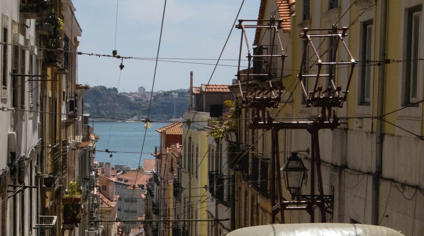 A narrow Lisbon street lined with pastel buildings and tram wires, sloping down toward terracotta rooftops and the Tagus River.
