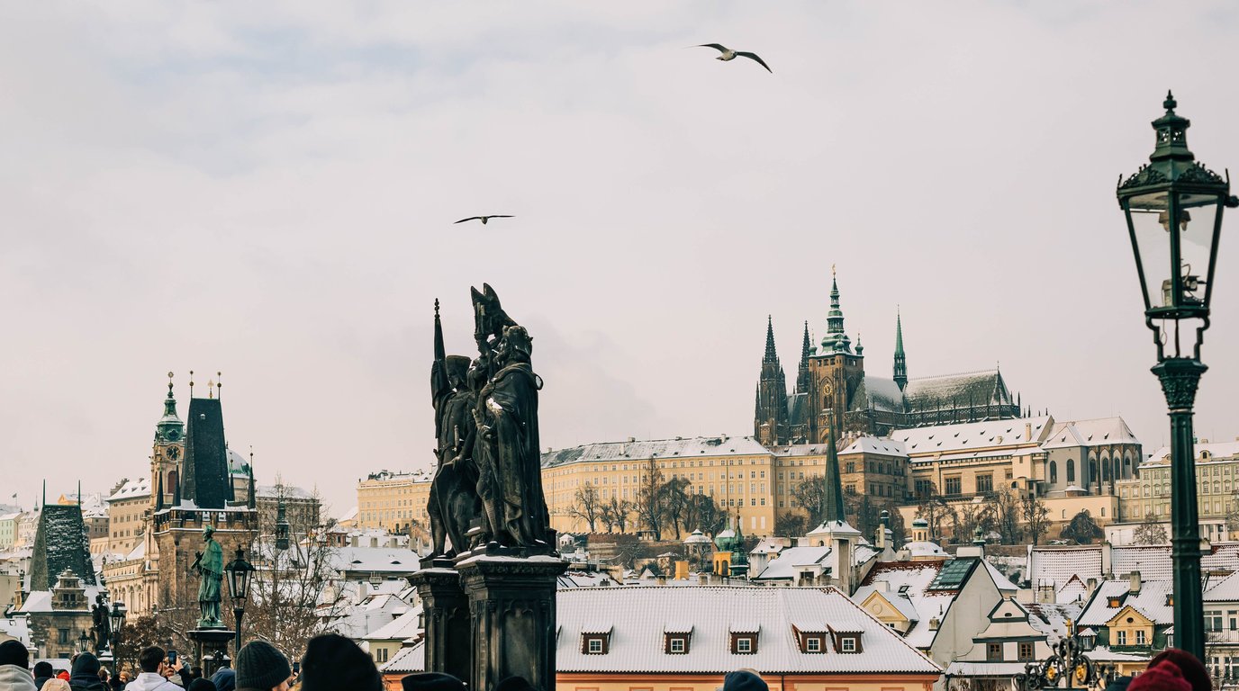 Snow-covered rooftops and Prague Castle rising above the city, seen from Charles Bridge on a winter day, with a bronze statue and an old street lamp in the foreground.