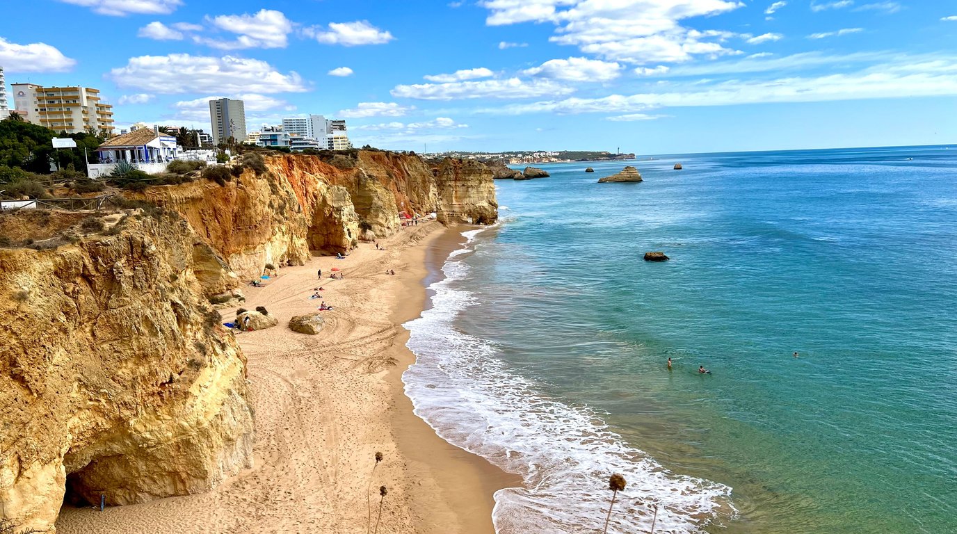 Golden sandstone cliffs line a wide sandy beach on Portugal's Algarve coast, with turquoise Atlantic water, a few swimmers, and clifftop hotels under a blue sky.