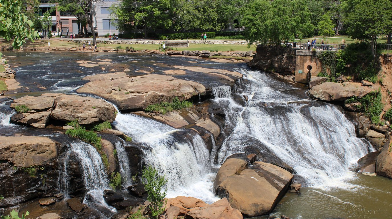 The Reedy River tumbling over wide rock ledges at Falls Park in downtown Greenville, South Carolina, with green lawns, trees and people walking along the park paths above.